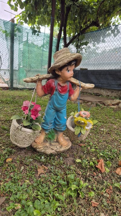 A garden statue of a young boy wearing a straw hat, a red shirt, and blue overalls stands in a yard. He is carrying a wooden pole across his shoulders, with a woven basket hanging from each end of the pole. The basket on his left holds pink artificial roses, and the basket on his right holds yellow and pink artificial flowers. The statue is made of a painted material and is placed on a roughly textured base. The background shows greenery, including tree branches with leaves and grapes overhead, a wire fence with a green mesh screen, and a dark tarp or fence. The ground is covered with grass, dirt, and fallen leaves. This image was taken in Doirentsi, Bulgaria.