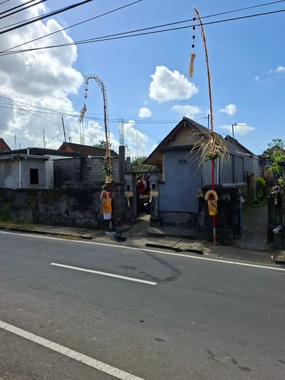 In Blahbatuh, Indonesia, on a bright, partly cloudy day, a street scene unfolds. The foreground is dominated by a paved road with white lane markings, suggesting it is a public thoroughfare. To the left of the road, a sidewalk runs alongside a wall, behind which are residential buildings. These structures are varied in design and condition, with some appearing aged and others under construction.
The most prominent features are two elaborately decorated poles, known as "penjor," which are traditional Balinese offerings. The first penjor, to the left, is tall and arched, adorned with woven palm leaves and small decorative elements. The second penjor, to the right, is a straighter, more slender pole, also decorated with palm leaves and smaller embellishments, including what appears to be a small figure or effigy attached. Both poles are adorned with red and yellow fabrics, indicating a festive or religious occasion.
The background reveals more of the residential area, with houses showing different architectural styles, some with tiled roofs and others with corrugated metal. Power lines crisscross the sky, a common sight in many urban and semi-urban environments. The overall impression is a snapshot of daily life in a Balinese village, marked by cultural traditions and the coexistence of older and newer structures.