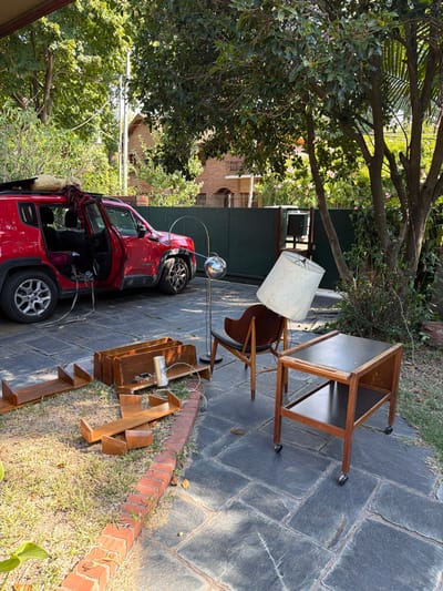 The scene depicts an outdoor residential area in San Isidro, Argentina, featuring a paved driveway or patio adjacent to a grassy section. A red Jeep Renegade SUV is parked on the dark flagstone pavement, with both its front and rear driver-side doors open. A light-colored, bundled object is secured by a red strap on the vehicle's roof rack. Cables or straps extend from the interior of the open doors to the ground.
Various furniture items are arranged on the pavement: a chrome arc floor lamp with a spherical, reflective shade stands upright. Next to it, a wooden armchair with a dark upholstered seat holds a large, white, conical lampshade resting askew on its back. To the right, a two-tiered wooden side table with a dark top surface is positioned on casters.
On the adjacent grassy area, bordered by a low line of red bricks, several disassembled wooden shelving components or bookends are scattered. A silver cylindrical object with white wiring is visible among these wooden pieces.
The background features dense green foliage from trees and shrubs, partially obscuring a multi-story brick residential building with a tiled roof, visible behind a dark green fence or wall. The lighting indicates daylight. No individuals are present in the image. The arrangement of objects suggests a state of staging for transport, loading, or unloading.