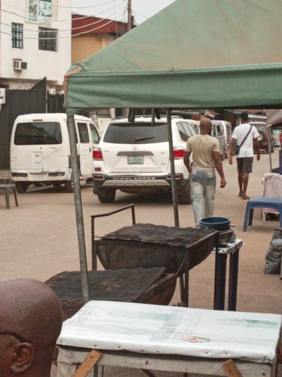 The image depicts a street scene in Owerri, Nigeria. In the background, there are several parked vehicles, including a white van and a white Toyota Highlander. Two men are walking away from the camera. One is bald and wearing a tan shirt and jeans, while the other is wearing a white shirt, dark shorts, and a black bag over his shoulder. In the foreground, there are charcoal grills, a table covered in white material, and a green awning overhead. The ground appears to be dirt or gravel. The building in the background appears to be two stories tall and has a faded advertisement on its side.