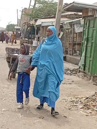This image depicts a street scene in the city of Gawuna, Nigeria. In the foreground, an adult female and a young male child walk from right to left on a dry, uneven dirt path. The female is dressed in a light blue full-body hijab covering her head and body, light blue trousers, dark socks, and grey slide sandals. Her eyes are visible, and she looks slightly to her right. She carries a dark bag on her left shoulder. The child, walking to her left, wears a grey hooded long-sleeve shirt with a graphic design, blue drawstring pants, and white sneakers. His hood is up, and he holds the woman's left hand.
The path is scattered with debris, including small sticks and refuse, particularly on the right side. To the left of the subjects, a two-wheeled cart carrying stacked cylindrical items is partially visible. The background consists of a row of modest market stalls or shops constructed with corrugated metal roofs and various materials for walls, some appearing weathered. A green metal door or shutter is visible on one structure on the right. Overhead, utility poles and electrical wires are present. Several blurred figures are visible walking further down the street in the distant background.