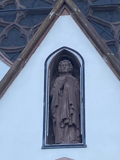 A stone statue is visible behind a black metal mesh screen in a rectangular opening in a white wall. The opening has a rounded top. The statue appears to be of a figure with curly hair, draped in fabric. To the left of the opening, there is a gothic-style stone framework with quatrefoil designs. The location is Bischheim, France.