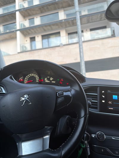 The image provides a view from inside a Peugeot vehicle, facing forward through the windshield. The black leather-wrapped steering wheel, featuring a silver Peugeot logo and an "AIRBAG" inscription, occupies the lower foreground. Controls are visible on the right spoke of the steering wheel. Behind it, the instrument cluster is illuminated, displaying a digital speed of "0," a trip meter of "208.9," and an odometer reading of "13562" kilometers. Red warning lights are visible on both sides of the cluster. To the right of the steering wheel, a set of keys, one with a green ribbon, hangs from the ignition. Further right, a dashboard-mounted infotainment screen shows "16°C" and active media playback with directional controls.
Beyond the windshield, a modern, light-colored multi-story building with dark-framed windows and balconies featuring transparent glass railings is visible under bright daytime lighting. A slender, metallic pole stands vertically in the mid-ground, partially obscuring the building. The scene depicts an urban environment characterized by contemporary architecture within the city of Vila Real, Portugal. No individuals, specific actions, or interactions are observable.
