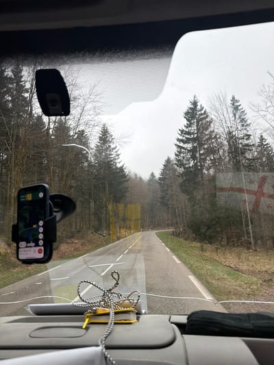 The image depicts a view from the interior of a vehicle looking out through the windshield onto a rural road in a forested area of Plaine, France. The two-lane asphalt road, which appears wet, features a broken white centerline and a solid white line marking the right shoulder. It curves gently to the right as it recedes into the distance. Both sides of the road are bordered by dense forest, consisting of a mix of tall evergreen conifers and leafless deciduous trees, indicative of late autumn or winter. The sky is overcast and gray.
Inside the vehicle, a smartphone displaying a navigation application with a visible blue route is mounted on a black suction-cup holder attached to the lower-left area of the windshield. The top of the dashboard is visible, on which a coiled black and white braided cable rests, next to a small yellow object. A dark, irregularly shaped housing is affixed to the upper central part of the windshield, likely for a rearview mirror or sensors. Visible reflections and distortions on the windshield include a white curved mark near the top left, and yellow rectangular patterns and a red cross-like image on the right side.