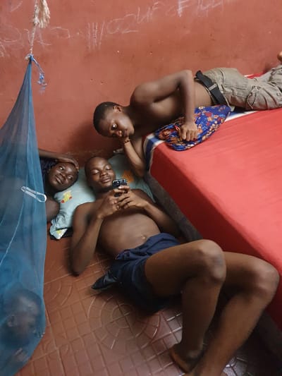 Four young individuals are seen relaxing on a red tiled floor in an indoor setting, viewed from a high angle. One person lies partly under a red mattress, wearing grey cargo pants and holding a blue patterned cloth. Adjacent to the mattress, another individual lies on their back, clad in dark shorts, with their knees bent.
Next to them, a third person lies on their back, holding a mobile phone over their chest, resting their head on a light blue pillow with a yellow pattern. A fourth individual lies on their stomach or side beside the third, also using the same pillow and looking upwards. A blue mosquito net hangs in the foreground, with part of another person's head faintly visible through it.
The overall atmosphere is one of casual relaxation. The red floor shows white chalk markings, including "2011" and tally marks. A rope with a blue tie, connected to the mosquito net, is also visible. The scene suggests a relaxed moment, possibly in the evening or night, within a modest home in Idah, Nigeria.
