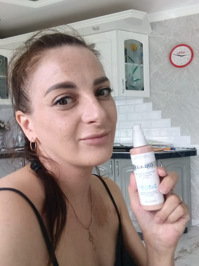The image shows a woman indoors, presumably in a kitchen, holding a bottle of "Enough Collagen Whitening Moisture Foundation" makeup. The background features white kitchen cabinets, a backsplash, a marble-patterned wall, and a red-rimmed clock. The woman has her hair pulled back in a ponytail and is wearing gold hoop earrings and a gold necklace. The precise location in Andreyevo, Russia, is not discernible from the image.