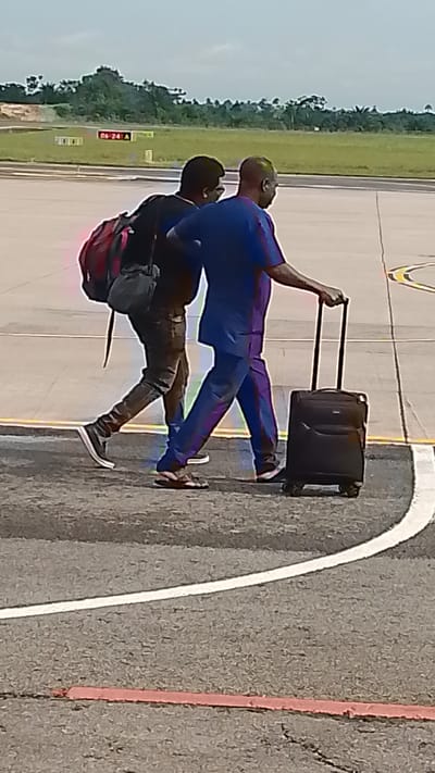 Two men are walking across an asphalt surface, possibly an airport tarmac, with a backdrop of lush green trees under a cloudy sky. The man in the foreground is wearing a blue dashiki and pants, and is pulling a black rolling suitcase. He has dark skin and a shaved head. The man behind him is wearing a black shirt and dark pants, and has a red backpack slung over his right shoulder. He is looking down at his feet. The scene appears to be an airport due to the markings on the ground, including white lines and a red strip. In the background, there are yellow signs with black text, one of which reads "06-24 A". The lighting suggests it is daytime. The overall mood is neutral, with no strong emotional cues. The activity is travel, as indicated by the luggage and the airport setting.