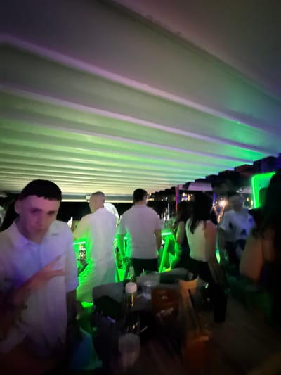 A group of people are gathered at an outdoor venue, possibly a bar or club, in Sozopol, Bulgaria. The scene is illuminated by vibrant green and purple lights, reflecting off a slatted ceiling. In the foreground, a young man in a white shirt is looking towards the camera, with his hand raised in a peace sign. Behind him, several other individuals are visible, some in white shirts, moving and interacting within the dimly lit space. Tables with drinks are present, suggesting a social gathering. The overall atmosphere appears to be lively and festive, characteristic of a nightlife setting.