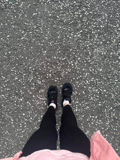 The image shows a top-down view of a person's legs and feet standing on asphalt in Magnitogorsk, Russia. The person is wearing black athletic shoes with pink accents, white socks, and black leggings. A pink jacket is visible at the bottom of the frame. The asphalt is dark gray and textured with small, light-colored stones. There are no other people or objects clearly visible in the scene. The lighting suggests it is daytime.