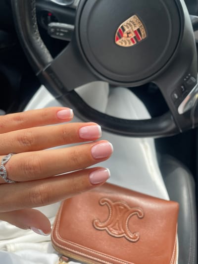 The image displays the interior of a vehicle, featuring a black leather steering wheel with a Porsche emblem centered on its hub, and a visible control stalk on the left. In the foreground, a person's left hand, with a light-to-medium skin tone, is prominently shown, exhibiting a French manicure with clear pinkish nail beds and distinct white tips. A silver-colored ring with an intertwined pattern is worn on the left ring finger. Below the hand, a light-colored fabric is visible. To the lower right, a brown leather clutch or wallet, embossed with the Celine Triomphe logo, rests on a dark surface, likely a car seat. The precise location within Santa Eulària Des Riu, Spain, cannot be determined from the visual information provided.