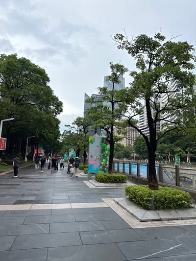 This image depicts a pedestrian walkway in Tianhe District, China, on an overcast day. The walkway is paved with large, dark gray rectangular stones, with a lighter colored stone stripe running down the center. Lush green trees line the left side of the walkway, casting shadows. On the right, a canal or waterway is visible, bordered by a metal railing. Beyond the railing, there are escalators leading down. In the background, modern glass and steel buildings rise against the cloudy sky. Several people are walking along the pathway. Some are dressed in casual attire, while others appear to be pushing strollers. There are also decorative elements on the right side of the walkway, including illuminated panels with green and colorful designs, and planters with manicured green bushes surrounding the bases of trees. A street lamp with a red digital display is visible on the left side of the walkway. The ground appears damp, suggesting recent rain.