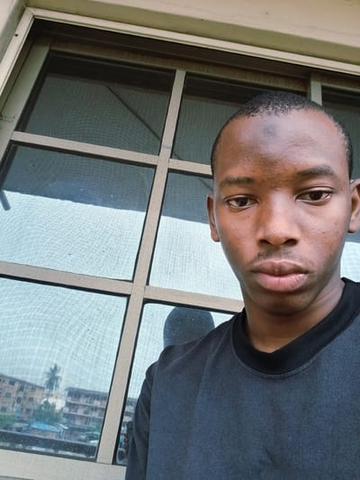 A young Black male is captured in a close-up selfie taken from a slightly low angle, positioned in front of a window. The background features a window with a grid of panes, some of which are covered by a fine mesh screen. Through the screen, buildings are visible, suggesting an urban environment. The lighting appears to be natural, coming from outside the window. The man is wearing a dark t-shirt. His expression is neutral, and he is looking directly at the camera. The overall scene suggests an indoor setting with a view of the outside.