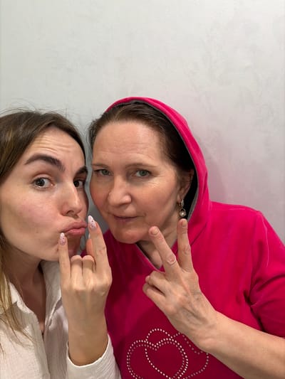 Two women pose for a selfie in what appears to be an indoor setting against a plain white wall. The woman on top wears a red hooded sweatshirt and displays a peace sign with her right hand. She has short brown hair, fair skin, and is wearing an earring with a pearl-like bead. The woman below has long brown hair and fair skin and appears to be making a duck face while holding up the "devil horns" gesture with her right hand. Her nails are painted white with red speckles. The scene provides no clues regarding the city of Neftekamsk or Russia.