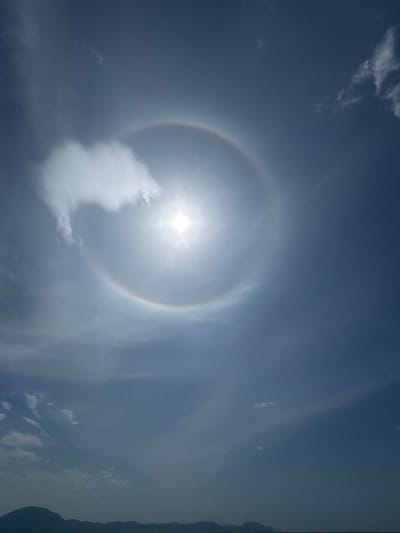 The image shows a bright sun with a halo effect in a clear blue sky. The halo is a circular arc of light with faint rainbow colors. Several wispy clouds are visible in the sky, with one larger, denser cloud on the left side of the sun. The bottom of the image features a dark, silhouetted horizon line, suggesting land or mountains against a faint body of water. The overall scene is a natural phenomenon occurring in an outdoor environment, possibly near a coast or large body of water given the horizon. The location is specified as Dong Ninh Hoa, Vietnam.