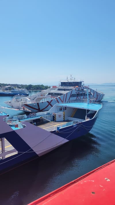 The image shows a sunny day with a clear blue sky over the port of Keramoti, Greece. Three ferries are docked or maneuvering in the calm, turquoise water. The closest ferry in the foreground is partially visible, with its dark blue and white patterned hull and a red railing. Behind it, a larger ferry, the "AMIRA," is prominently featured, displaying a distinctive red and white zigzag pattern on its hull and upper decks. Its bow is angled towards the viewer, and people can be seen on its deck. Further in the background, another ferry, identified by the text "THASSOS SEAWAYS" on its side, is also docked, with buildings and lush green trees visible on the shoreline behind it. The scene captures a typical day at this coastal transportation hub.