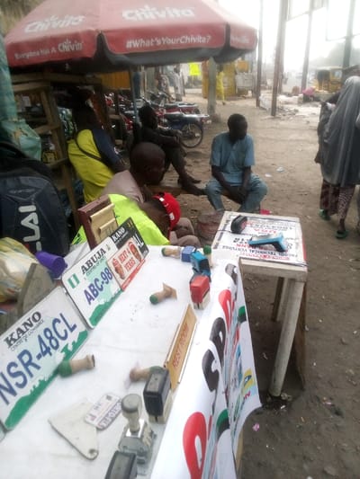 The scene depicts an outdoor street vendor stall in Sabon Gari East, Nigeria. Underneath a red Chivita-branded umbrella, various individuals are present. A man in a light blue tunic is seated on a low stool to the right of a white table display, looking towards the right. Another individual in a light brown shirt and red cap is positioned over the table, obscured, suggesting leaning or sleeping. A third person, wearing a yellow and blue shirt, is seated or leaning near a black backpack to the left. Behind them, another man sits on a bench in darker attire, and multiple motorcycles are parked.

The white table, supported by wooden trestles, serves as the primary display. On its surface are two Nigerian vehicle license plates: one green and white for "ABUJA ABC-98 FEDERAL REPUBLIC" and another white and green for "KANO NSR-48CL". Numerous hand stamps, including self-inking and traditional wooden-handled types, are scattered across the table. A small "REST ROOM" sign and a black electronic device are also present. The front of the table is draped with a white banner displaying text and logos, including "FACOOL TECHNICAL VENTURES" and a "UBA" bank logo. The ground is unpaved, appearing as dirt or loose earth. In the background, an unpaved road extends, populated by additional individuals, structures, and a tricycle taxi, under hazy daylight conditions. A person in light-colored religious attire walks away from the camera on the far right.