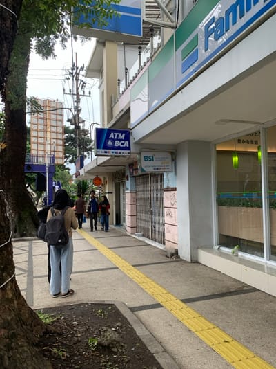 The image shows a sidewalk in front of a building in Malang, Indonesia, located at coordinates -7.98 latitude and 112.63 longitude. There are several people walking on the sidewalk, including a woman in a hijab carrying a backpack and three other people walking ahead of her. The building has a sign for "ATM BCA" and "BSI" with an ATM sign below it. The building has large windows, with an interior visible. The sidewalk is paved with concrete and has a yellow tactile paving strip running down the center. The building has a painted, pink facade. A large tree is visible in the left foreground. There is a blue and green sign above the building with Chinese characters on it. The building has metal security gates, which are closed. There is a green bush outside the window. The image is taken at a slight angle, looking down the sidewalk.