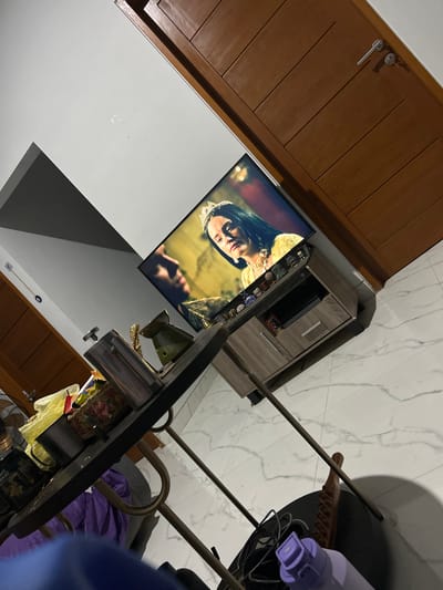 The image depicts an indoor scene in what appears to be a living room or entertainment area, with the camera angled from a high perspective looking down. The primary subjects are two people shown on a television screen, engaged in a scene that seems dramatic or emotional, with one person's face close to the other. The lighting on the television suggests it is evening or the room's lighting is dimmed for viewing.
In the foreground, a dark-colored table or desk is visible, with various objects placed on it, including decorative items and a yellow plastic bag. A purple bottle or container is also present on a surface to the right. Behind the television, a light-colored entertainment unit with drawers and shelves is positioned against a wall.
The room has a marble-tiled floor, reflecting light from above. To the left, a wooden cabinet or door with horizontal panels is visible. The overall atmosphere seems relaxed and domestic, likely a home environment in Sukawati, Indonesia, given the location context. There is no discernible text within the image. The composition of the shot, with its tilted angle, suggests it was taken by someone standing or sitting nearby, looking down at the scene.