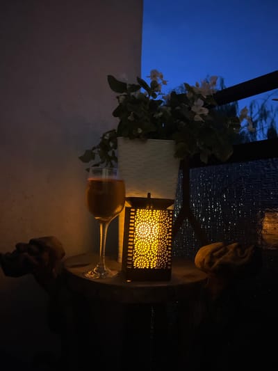 This image captures a serene evening scene on a balcony or patio, possibly in the city of Kleosin, Poland. The setting is dimly lit by the warm glow of a decorative lantern, casting intricate patterns on its surface. A tall, slender glass filled with a golden beverage, likely champagne or a similar sparkling drink, stands next to the lantern.
Behind the glass and lantern, a potted plant with delicate white flowers is visible, its leaves creating a soft silhouette against the darkening sky. The sky itself is a deep, rich blue, transitioning to darkness, suggesting twilight or early night.
The table surface appears to be made of wood, with a weathered or textured appearance. To the left and right of the central arrangement are two amorphous, fabric-like objects, their forms indistinct in the low light. These could be cushions, throws, or other decorative elements.
In the background, a dark railing and a textured surface, possibly a wall or window covering, can be observed. The overall atmosphere is one of peaceful relaxation and quiet contemplation, enhanced by the soft lighting and the evening sky. There are no people visible in the image.