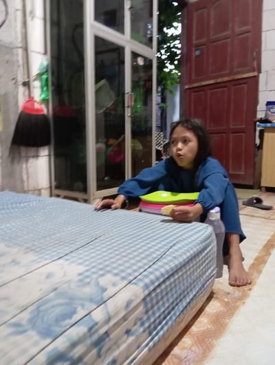 A child in a blue long-sleeved shirt is seen holding a stack of colorful bowls in what appears to be a domestic interior. The child is positioned near a mattress with a blue and white patterned cover. In the background, there is a glass-fronted cabinet or window, and a red door. A broom is standing in the bottom left of the image. The floor near the top right is tiled with a geometric pattern. The location is indicated as Baron, Indonesia, though the specific scene offers no distinctive landmarks to confirm this.