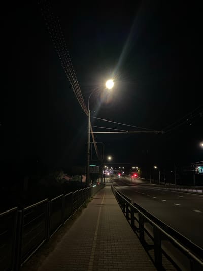 The image depicts a nighttime view of an urban road in Brest, Belarus. A brightly lit street lamp casts a strong glare in the upper center of the frame. To the left of the road is a pedestrian walkway paved with interlocking bricks, bordered by a metal fence. Overhead, power lines are visible, strung with decorative lights. The road itself is dark asphalt, with white lane markings. Traffic lights are visible in the distance, with at least one showing red. Buildings and trees can be seen on the left side of the road, and a billboard is present on the right. The overall scene suggests a quiet, possibly deserted, urban environment after dark.