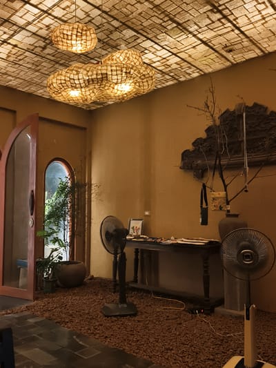 The image depicts an indoor scene, possibly a cafe or restaurant, with a distinctly rustic and natural aesthetic. The ceiling is adorned with a grid-like pattern, illuminated by three large, irregularly shaped pendant lamps made of what appears to be woven natural material. These lamps cast a warm, diffused glow, contributing to the overall ambiance.
The walls are a textured, earthy brown, providing a backdrop for several decorative elements. On the right side of the image, a dark, intricate carved wooden panel is mounted on the wall. In front of this panel, a bare branch with sparse leaves is placed in a large, dark vase, adorned with two small bags hanging from its twigs.
The floor is a combination of dark, rectangular tiles in the foreground and a layer of reddish-brown gravel covering most of the floor area. Two black oscillating fans are present; one is a traditional pedestal fan to the left of a dark wooden table, and another is a more modern, sleek fan on the right. The table is set with various items, including papers, a small framed picture, and what seem to be office supplies.
To the left, an arched doorway, partially ajar, reveals another archway beyond, through which glimpses of greenery from an indoor plant can be seen. A large, round planter sits near this doorway. The lighting suggests an interior space with artificial illumination, possibly supplemented by natural light from the exterior visible through the doorways. The overall impression is one of a thoughtfully designed space aiming for a natural, handcrafted, and slightly bohemian feel. The location in Hoa Lư Ward, Vietnam, suggests a potential Vietnamese influence on the decor and materials used.