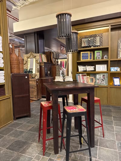 The image depicts an indoor scene, likely a retail environment, featuring a rustic-style dining set and shelving units. In the foreground, a dark wooden table stands on a tiled floor, accompanied by three stools: two red and one black. The red stools have a Coca-Cola-themed print on their seats, while the black stool has a similar design. Behind the table, a dark wooden cabinet is visible, contrasting with the lighter wood of the shelving units behind it.
The shelving units display a variety of decorative items, including framed pictures, photo frames, decorative birdhouses, and other assorted knick-knacks. A large, ornate mirror with a gold frame is placed on top of a wooden chest of drawers to the left of the table. Woven baskets and stacks of folded towels are also present in the composition. The ceiling features exposed ductwork and lighting fixtures, typical of a commercial space.
The scene does not contain any individuals. The overall aesthetic suggests a bohemian or eclectic decor style.
While this image depicts a store interior, its specific location in Kuala Lumpur, Malaysia, cannot be determined from the visual information alone.