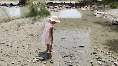 A young girl wearing a light pink dress and a straw hat stands on a muddy, rocky shore next to a shallow, clear body of water. The girl is facing away from the camera, looking down into the water. Her sandals are visible on her feet. The environment appears to be a natural setting with a river or stream, rocks, and vegetation. There is no indication of the city of Taldykorgan, Kazakhstan, in the image.