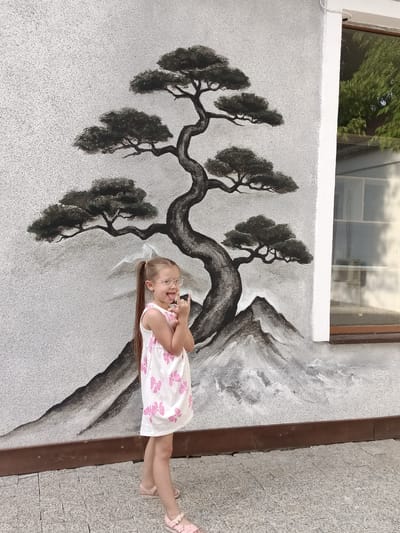 A young girl with blonde hair in a ponytail, wearing a white dress with pink flowers and pink sandals, stands in front of a wall mural of a stylized tree and mountains. She is sticking her tongue out and making a rock-and-roll hand gesture. The mural is painted in black and gray tones on a textured, light-colored wall. To the right of the mural is a window with reflections of trees. The ground is paved with light gray stones. The setting appears to be outdoors, likely in a public space or a building's exterior. The lighting suggests it is daytime.