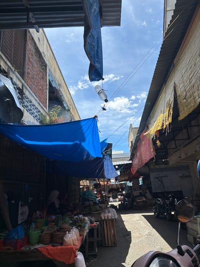 A street market is depicted in Bireuen, Indonesia. The scene is characterized by a narrow alleyway lined with market stalls covered by tarps and corrugated metal roofs. On the left, a stall displays various produce, including vegetables and fruits, as well as pottery and sacks of rice. Two women, one wearing a hijab, are seated behind the stall. In the background, several motorcycles are parked along the right side of the alley. The sky above is blue with scattered clouds. The overall impression is one of a busy, open-air market under a bright, sunny sky.