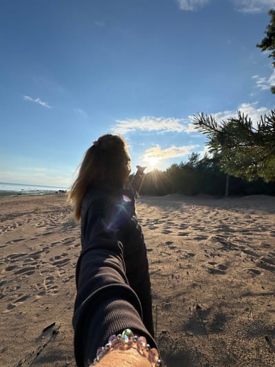 This is a photo taken from a person's perspective at a beach. The person is lying on their side on the sand, looking towards the sky and the ocean. The sun is setting or rising, casting a warm glow on the scene and creating lens flare. Pine trees are visible on the left side of the frame, indicating a natural setting. The sky is a clear blue with some scattered clouds. The sand is textured with footprints. The person is wearing a dark-colored top and a beaded bracelet on their wrist. The overall mood is serene and peaceful, suggesting a moment of relaxation and appreciation of nature. The location is identified as Shepelevo, Russia.