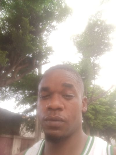The image is a selfie taken outdoors in Gwarinpa, Nigeria. A young Black male, with short hair and dark skin, is in the foreground, looking directly at the camera with a neutral expression. He is wearing a white tank top with green trim. Behind him, large trees with green foliage are visible, suggesting a park or a residential area with significant greenery. The sky is bright, indicating it is daytime, though there appears to be some overexposure. A portion of a building with a patterned facade is also visible in the lower left corner of the frame. The overall scene suggests a casual, outdoor setting within the city.