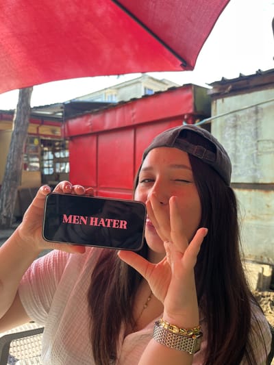 A person is depicted outdoors, holding a smartphone to display its screen. The individual appears to be female, wearing a dark baseball cap backward and a light-colored, possibly white, textured shirt. Long, dark hair cascades around her shoulders. She wears a watch with a silver and gold band, and a gold-colored bracelet on her left wrist. Her right hand partially covers her face, with one eye visible, looking down towards the phone.

The smartphone, held in her left hand, displays the words "MEN HATER" in prominent pink capital letters against a black background.

The setting appears to be an outdoor area, possibly a cafe or market street in Karnobat, Bulgaria. Large red awnings or umbrellas are visible on the left and in the background, providing shade. Behind the person, aged structures with what look like wooden or metal elements and some windows can be seen. The bright lighting suggests it is daytime. The overall impression is of a casual moment where the person is making a playful or defiant statement by displaying the text on her phone.