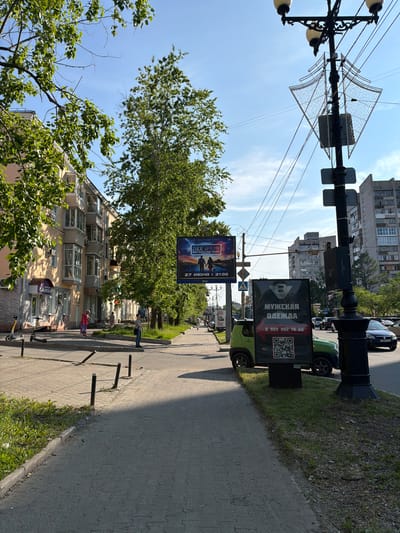This is a street-level, eye-level, medium-range, medium-shot, full-color, daytime photograph taken in Khabarovsk, Russia. The image captures a sidewalk scene with a street and buildings in the background.

In the foreground, a paved sidewalk runs along a grassy area. A street with parked cars and moving traffic is visible to the right. On the left side of the sidewalk, there is a building with light-colored walls and several balconies. In front of the building, there are two electric scooters parked.

Further down the sidewalk, a large billboard is displayed, featuring an advertisement for an event with text in Russian, including a date and time. Near this billboard, a small green vehicle is parked with a black advertising display attached to its rear, also featuring Russian text and a QR code. A lamppost with multiple lights and wires is present in the middle ground, with a decorative element attached to it.

In the background, there are several multi-story buildings, characteristic of urban architecture in Khabarovsk. The sky is blue with some scattered clouds. The overall environment appears to be a typical city street in a temperate climate during the day.

There are a few people visible walking on the sidewalk, dressed in casual clothing, suggesting normal daily activity. The scene is well-lit by sunlight, casting shadows on the ground. The presence of the electric scooters and the advertising displays indicates a commercial and urban setting.