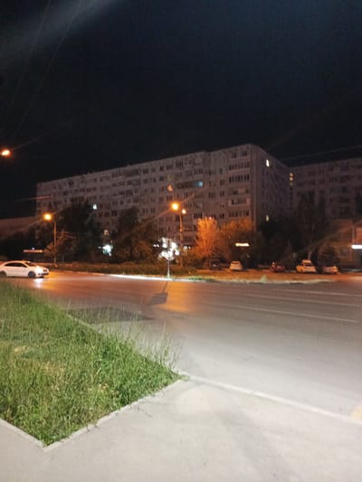 A night-time outdoor shot captures a long, multi-story apartment building in Volzhsky, Russia, illuminated by streetlights and lights from within some apartments. In the foreground, a paved road runs horizontally, bordered by a grassy median strip and a sidewalk. A white car is seen driving on the road, its headlights illuminating the asphalt. Several other cars are parked along the street in the middle ground. The sky is dark and clear, suggesting it is nighttime. The scene depicts a typical urban environment at night.