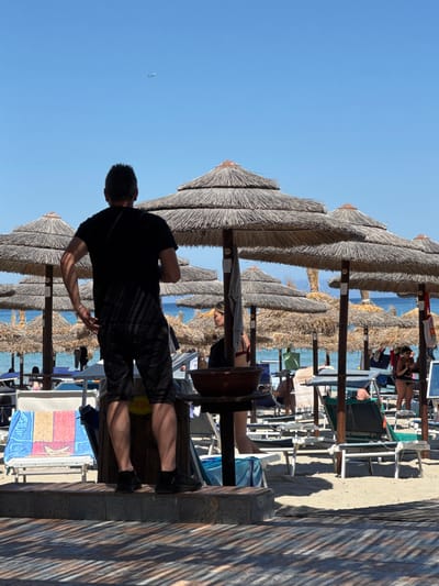 The image depicts a sun-drenched beach scene in Isola delle Femmine, Italy, with a clear blue sky. In the foreground, a man stands with his back to the camera, wearing a black t-shirt and shorts. He is positioned on a raised wooden platform with tiled accents, casting a shadow from the overhead thatched umbrellas. Behind him, several straw umbrellas are lined up on the sandy beach, with lounge chairs scattered beneath them. The turquoise sea is visible in the background, with a distant airplane visible in the sky. Several other beachgoers are present, some seated on lounge chairs and others walking among the umbrellas. A woman in a bikini is visible in the middle ground, and a folded towel with a colorful pattern lies on a lounge chair to the left. The overall impression is a typical summer day at a seaside resort.