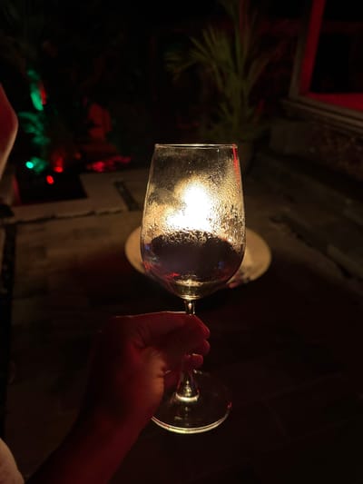 A hand holds a condensation-covered wine glass in a dim, indoor setting in Ungasan, Indonesia. Red and green lights are visible in the background, suggesting a holiday or decorative lighting setup. A white object, possibly a plate or coaster, sits behind the glass. The hand appears to be gripping the stem of the wine glass. The environment is dark, with hints of furniture or architectural elements in the periphery.