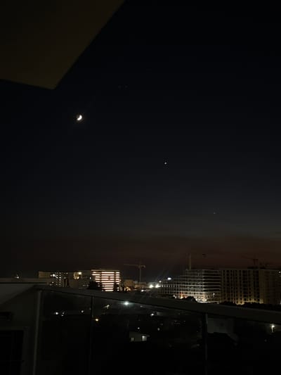 The image depicts a nighttime cityscape with a crescent moon and two visible stars in the dark sky. In the foreground, a balcony railing is visible, suggesting the viewpoint is from a high-rise building. The background features several multi-story buildings, some of which are illuminated, indicating urban development. Construction cranes are also present, suggesting ongoing building projects. The sky transitions from a deep dark blue to a faint reddish hue near the horizon. The overall scene suggests an evening view over the city of Šušanj, Montenegro.