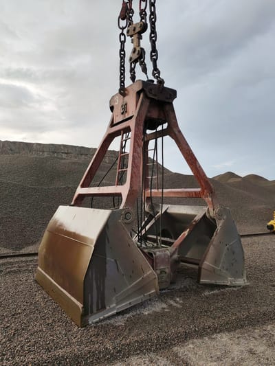 A large, rusty orange grab bucket is suspended by chains above a pile of dark gray gravel. The bucket has two large, shovel-like scoops, both open and appearing wet. Behind the bucket, a vast landscape of gravel extends into the distance, with rolling hills of the same material. The sky above is overcast with gray clouds. In the far right background, a small portion of a yellow vehicle is visible. The scene appears to be an industrial area or a construction site.