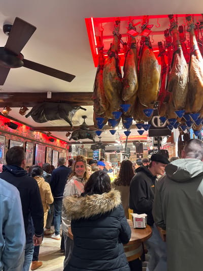 The image shows a crowded restaurant in Donostia, Spain, located at latitude 43.324 and longitude -1.986. The restaurant has a ceiling adorned with hanging cured hams, a large, black, bull-shaped sculpture hangs from the ceiling near the back of the restaurant, and a ceiling fan is in the foreground. There are approximately 20 people in the restaurant, including men, women, and children. The majority of the people are standing, and some are seated at tables. The restaurant is dimly lit, and the walls are adorned with posters and artwork. There is a wooden bar near the front of the restaurant.  A young woman in a light-colored jacket stands near the front, looking at the camera, and a younger woman in a black jacket with a fur collar is standing to her right, looking down. Two men, one with dark hair and wearing a black jacket, and the other with light hair and wearing a grey jacket, are standing next to the young woman in the light-colored jacket. A man with brown hair and wearing a black beanie is standing behind the woman in the black jacket.  The image was likely taken using a smartphone camera held in the hands of an individual standing in the restaurant, which is why the image includes the ceiling fan in the foreground.