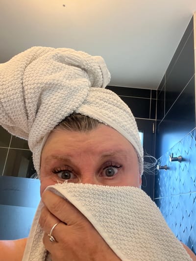 A person, likely a woman, is seen from a close-up, high-angle perspective. She has a white towel wrapped around her head like a turban and is holding part of the towel to her face. Her gray eyes are visible, and she wears a ring on her finger. The background suggests a bathroom setting with black and white tile. The location appears to be a bathroom in a private residence. The city is not relevant.