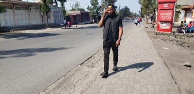 A man in a black vest, black jeans, and a yellow cap walks along a sidewalk, talking on a cell phone. He casts a shadow on the sidewalk. A red mailbox covered in graffiti stands nearby. The scene is outdoors, likely in a small town, with a paved road, buildings, and other people in the background. The weather appears sunny and clear.