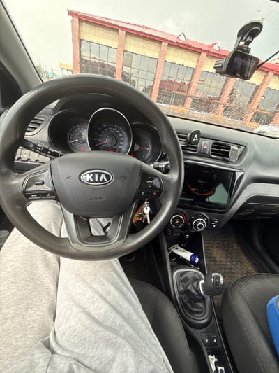 The image provides an interior view of a car, likely a Kia, from the driver's perspective. The dashboard is dominated by a steering wheel with the Kia logo prominently displayed, and a digital instrument cluster showing speed and RPM readings. To the right of the steering wheel, a touchscreen infotainment system is visible, displaying "Моя дочка" (My daughter) and some graphical elements. Below the screen are climate control knobs. A manual gear shifter with R, 1, 3, and 5 indicated is also present, suggesting a manual transmission. A dashcam is mounted on the windshield, its screen showing a reflection of a building with red and yellow facades and numerous windows. The weather appears overcast, with some water droplets on the windshield, creating distorted reflections. The car interior has dark grey plastics and black fabric seats. A person's legs, clad in light grey sweatpants, are visible in the foreground, indicating the presence of the driver or a passenger. The scene is set inside a vehicle, likely parked or driving in a location that has such buildings visible. The overall impression is a candid snapshot of a car's interior.