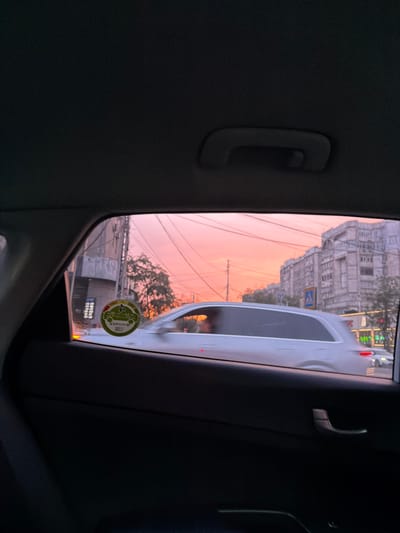 The image, taken from inside a vehicle, captures a view through the side window during what appears to be sunset. The sky is painted in hues of soft orange and pink, suggesting the golden hour. In the distance, an urban landscape is visible, characterized by multi-story buildings with traditional architecture, possibly in Bishkek, Kyrgyzstan. A white car is in motion or parked, partially obscuring the view of the buildings and the sky.
Streetlights and power lines crisscross the scene, adding to the urban setting. On the window, a circular sticker with green and yellow elements is visible, possibly indicating vehicle registration or a local decal. The sticker contains some text and a graphic that appears to be a stylized car or leaf. The interior of the vehicle is dark, with a prominent handle visible on the left side, suggesting the perspective of a passenger. The overall mood is tranquil, with the warm colors of the sunset creating a serene atmosphere against the backdrop of the city. The scene is likely taking place in the late afternoon or early evening as daylight fades.