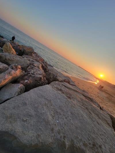 The image shows a sunset scene at the Quarteira beach in Portugal. A person is seen standing on the rocky shore, possibly fishing, with the ocean stretching out to the horizon. The sun is setting, casting a warm orange glow across the sky and reflecting on the water. Further down the beach, a small group of people are visible walking. The foreground is dominated by large, textured rocks, and the background shows the sandy beach and the calm sea. The sky transitions from a pale blue at the top to hues of orange and yellow near the horizon.