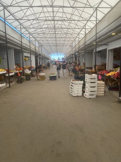 A covered outdoor market in Sevastopol, Ukraine, features rows of stalls selling produce. The market is covered by a white translucent roof supported by a metal frame. Sunlight filters through the roof, illuminating the scene. A paved walkway runs through the center of the market, flanked by stalls on either side. The stalls are filled with a variety of fruits and vegetables, including bananas, apples, tomatoes, and oranges. Several people are browsing the stalls or walking along the walkway. Some people are wearing casual clothing, such as t-shirts and shorts. A few people appear to be vendors, arranging their merchandise. Boxes and crates are stacked in front of some stalls, likely containing produce or packaging. The overall atmosphere is one of a busy, open-air market on a clear day.