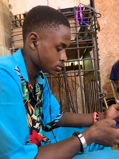 A person with dark skin and short, dark hair is wearing a light blue shirt with a floral patterned collar and has earbuds in their ears. They are standing in front of a barred structure that appears to be a window or gate. The bars are dark, and there is a padlock hanging from them. The ground is tiled with a brown and beige patterned tile. There is evidence of debris and clutter behind the bars. The setting appears to be indoors, near an exterior opening, and possibly within the city of Akpaka Forest Reserve, Nigeria. The person is looking down, possibly at something they are holding or working on. There are bracelets on their wrist.