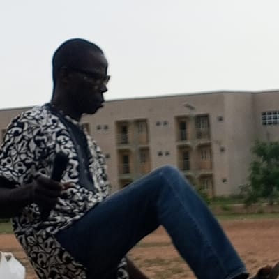 The image shows a Black man in a patterned shirt and blue jeans, sitting outdoors with his left leg raised. He is wearing glasses and holding a dark object, possibly a microphone, in his left hand. In the background, a light brown building with multiple windows is visible, along with some trees and a cloudy sky. The ground appears to be dirt. The scene is likely located in Barden Yana, Nigeria, given the style of the building and vegetation.