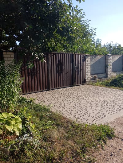 The image depicts an exterior view of a property entrance in Sozonivka, Ukraine, on a bright, clear day. The scene is dominated by a large, dark brown metal gate, which appears to be a double gate, closed. The gate has vertical ribbed panels and decorative scrollwork on each leaf. To the left of the gate, lush green foliage, including large hosta leaves, grows densely. Beyond the gate and to the right, a brick wall with concrete panels forms the boundary of the property. The ground in front of the gate is paved with light-colored interlocking bricks, forming a driveway or pathway. Sunlight casts distinct shadows, indicating it is likely midday or early afternoon. The overall impression is one of a well-maintained, private residential entrance under a clear sky, with vegetation adding a natural element to the constructed elements. There is no visible text in the image.