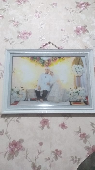 A white-framed photograph hangs on a light-colored wall covered in floral wallpaper featuring pink and purple blossoms with green foliage. The rectangular frame has an inner silver border and is suspended by a dark string from a single point at the top center.
The photograph within the frame depicts two individuals, a male and a female, in formal attire, positioned in a decorated setting. The male, wearing a white long-sleeved tunic, dark trousers, and a white skullcap, is seated on a light-colored, upholstered bench. The female, dressed in a white bridal gown and a white hijab, is positioned next to him, her arm linked with his. Both subjects face forward, looking towards the viewer. The background of the photograph is a softly lit, pale yellow backdrop. White floral arrangements serve as decor, including a large cluster on the left, a tall arrangement on a stand on the right, and a lower floral cluster on the right. A small red mat is visible beneath the floral arrangement on the left.
The specific city of Ilie, Indonesia, is not identifiable from the visual content of this image.